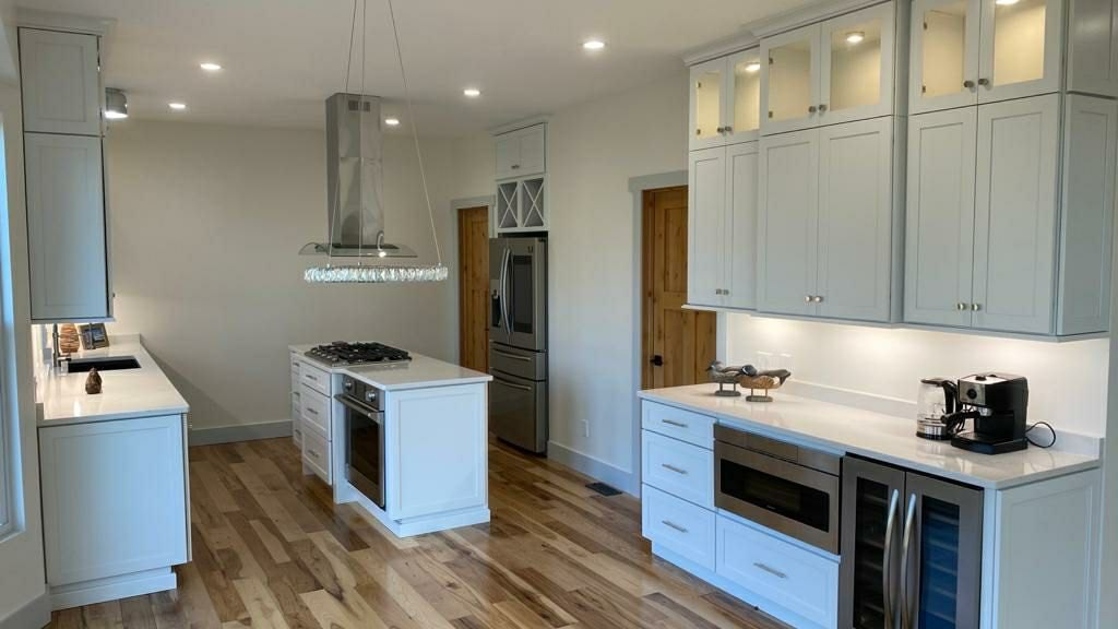 Kitchen with pale grey Shaker cabinetry, lit glass-front uppers, white quartz countertops and rustic hickory plank flooring